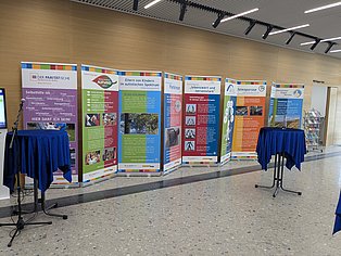 Banner verschiedener Selbsthilfegruppen stehen entlang einer Wand. Zwei Stehtische stehe davor.