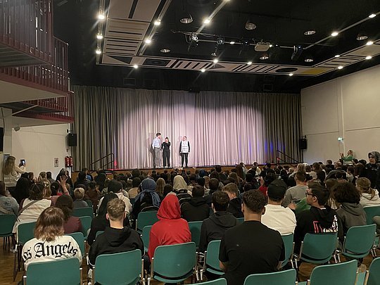 Saal mit Publikum und 3 Personen auf der Bühne